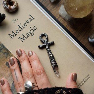 Può includere: Un ciondolo ankh in metallo scuro con una superficie ruvida e testurizzata e una punta di cristallo trasparente. Il ciondolo poggia su un libro intitolato IV. Medieval Magic, circondato da vari cristalli e una sfera di vetro su legno.