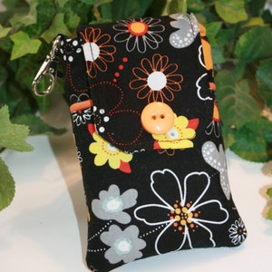 Peut inclure: Une pochette en tissu noir avec un motif floral orange, jaune, gris et blanc. La pochette a une fermeture à rabat avec un bouton orange et un fermoir argenté. Le motif présente de grandes et petites fleurs.