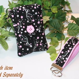 Peut inclure: Un étui de téléphone et une dragonne en tissu noir avec un motif floral de branches blanches et de fleurs roses. L'étui de téléphone a une fermeture à bouton rose et un fermoir argenté. La dragonne a un intérieur rose.