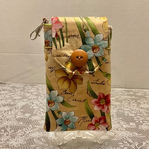 May include: A beige floral print phone case with a flap closure and a large wooden button. The case features a pattern of colorful flowers and handwritten script. It has a silver-tone clasp and is displayed on a clear stand.