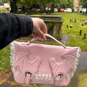 May include: Pale pink satchel bag with bat wing detailing and white lace trim. The bag has a top handle, a flap closure with two buckles, and a structured design. The bat wings are stitched onto the front.