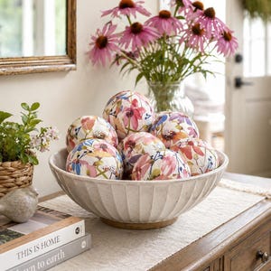 May include: A decorative bowl filled with floral-patterned spheres. The bowl is off-white with vertical ridges. A bouquet of pink coneflowers sits in a glass vase behind the bowl. The table also has books with the titles "THIS IS HOME" and "down to earth".