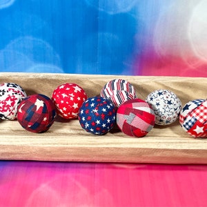 May include: A wooden bowl filled with nine fabric-covered balls in red, white, and blue patriotic patterns. The balls feature stars, stripes, and plaid designs.