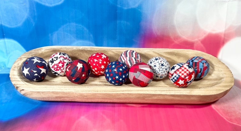 May include: A wooden bowl filled with nine fabric-covered balls in red, white, and blue patriotic patterns. The balls feature stars, stripes, and plaid designs.