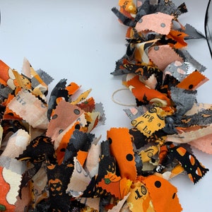 May include: A festive Halloween garland made of fabric scraps in orange, black, gray, and peach. The fabric pieces are cut into strips and tied together, featuring patterns like pumpkins, cats, and polka dots. A decorative accent for the spooky season.