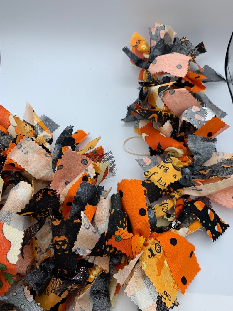 May include: A festive Halloween garland made of fabric scraps in orange, black, gray, and peach. The fabric pieces are cut into strips and tied together, featuring patterns like pumpkins, cats, and polka dots. A decorative accent for the spooky season.