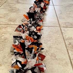 May include: A long, handmade Halloween garland made of fabric strips in black, orange, and white. The fabric features Halloween-themed patterns like ghosts, bats, and pumpkins. The garland is laid out on a tiled floor.
