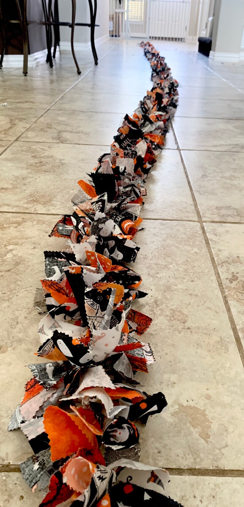 May include: A long, handmade Halloween garland made of fabric strips in black, orange, and white. The fabric features Halloween-themed patterns like ghosts, bats, and pumpkins. The garland is laid out on a tiled floor.