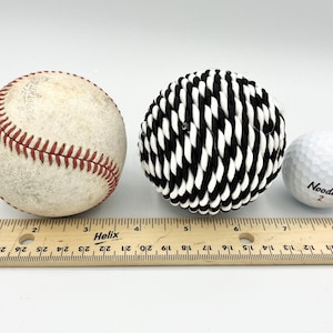 May include: Three balls of different sizes and textures: a white baseball with red stitching, a black and white rope ball, and a white golf ball with the word "Noodle" and the number "2" printed on it. A wooden ruler is in the foreground.