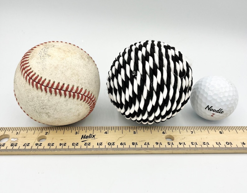 May include: Three balls of different sizes and textures: a white baseball with red stitching, a black and white rope ball, and a white golf ball with the word "Noodle" and the number "2" printed on it. A wooden ruler is in the foreground.