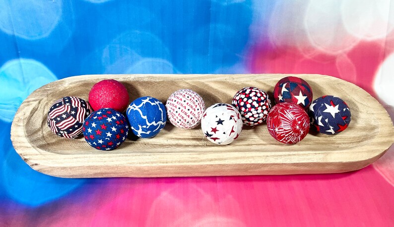 May include: A wooden bowl filled with red, white, and blue fabric balls. The balls have various patriotic patterns, including stars, stripes, and fireworks.