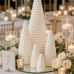 May include: A set of five cone-shaped decorative ornaments covered in glossy white faux pearls of varying sizes. The ornaments are arranged in a cluster on a circular mirror base, surrounded by white roses, greenery, and glass candle holders.