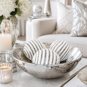May include: A large, round, silver-toned hammered metal bowl containing three decorative spheres. Each sphere is constructed from white rope coiled in a circular pattern, with thin, sparkling silver rhinestone bands embedded between the rope rows.