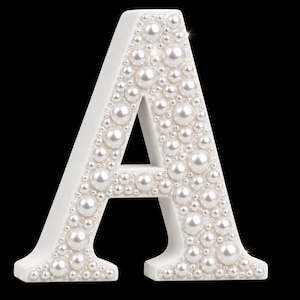 Peut inclure: Un objet décoratif en forme de lettre A blanche, dont la face avant est densément recouverte de perles artificielles blanches, rondes et brillantes, de tailles variées. La lettre possède une structure de base blanche et unie.