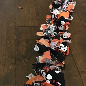 May include: A Halloween-themed garland made of orange, black, and white fabric scraps. The garland features various patterns, including polka dots, spiders, and the word "boo". The garland is laid out on a wooden surface.