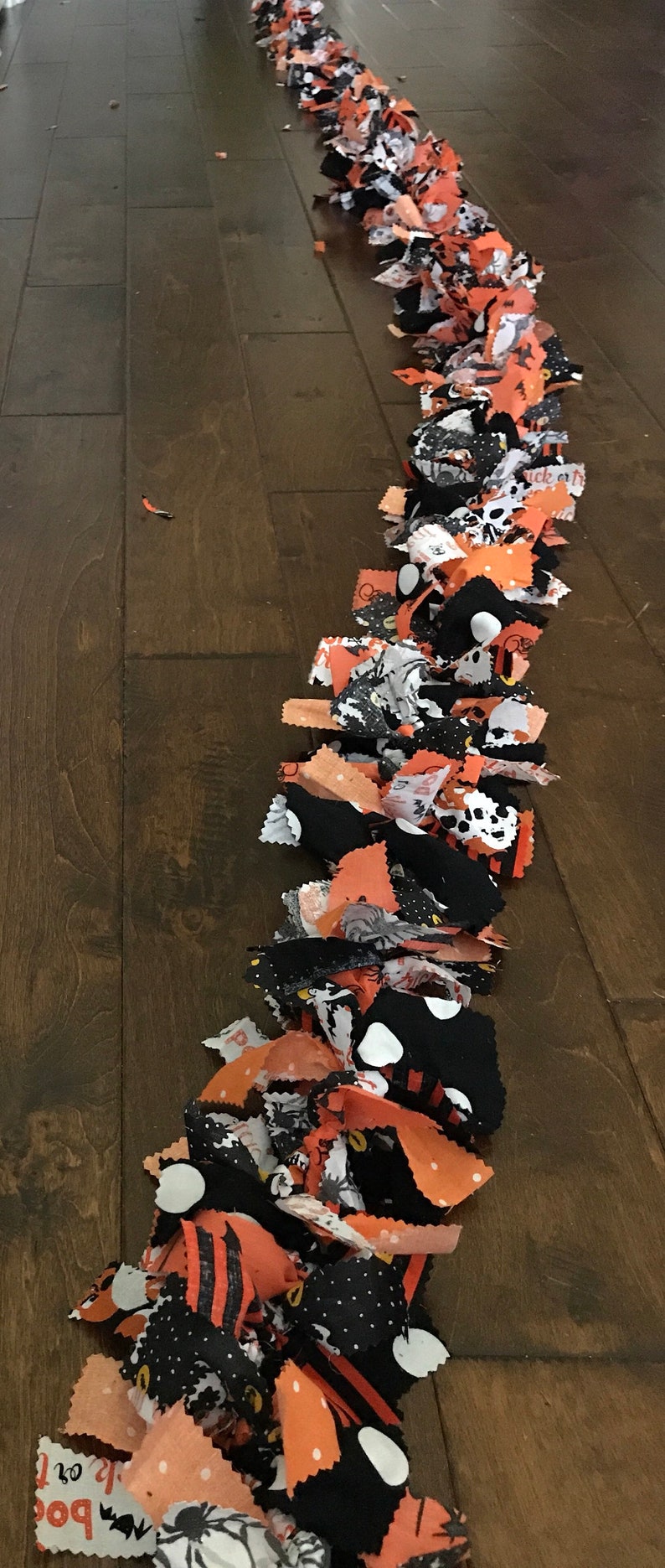 May include: A Halloween-themed garland made of orange, black, and white fabric scraps. The garland features various patterns, including polka dots, spiders, and the word "boo". The garland is laid out on a wooden surface.