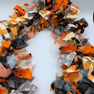 May include: A festive Halloween garland made of fabric strips in orange, black, gray, and cream. The fabric features pumpkin, candy corn, and plaid patterns. This handmade garland is perfect for seasonal home decor.
