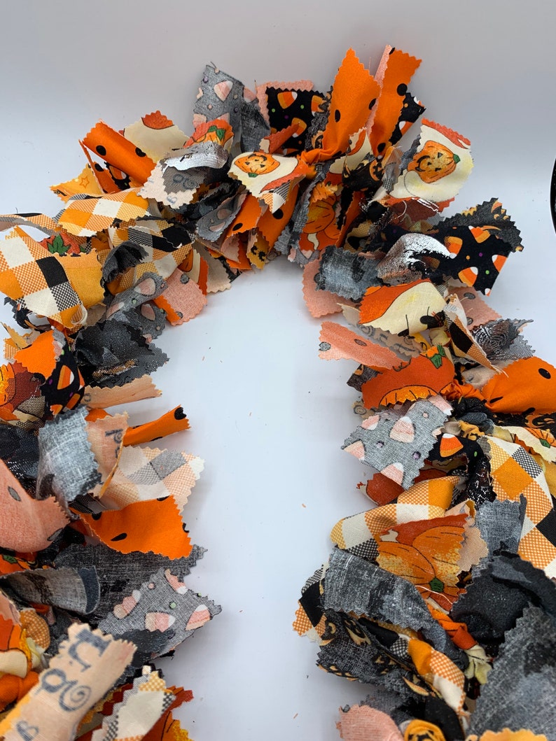 May include: A festive Halloween garland made of fabric strips in orange, black, gray, and cream. The fabric features pumpkin, candy corn, and plaid patterns. This handmade garland is perfect for seasonal home decor.