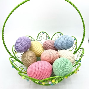 May include: A green wire basket with a handle filled with colorful yarn Easter eggs. The eggs are in shades of pink, blue, yellow, and brown.