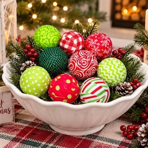 May include: A white scalloped ceramic bowl filled with assorted fabric-covered Christmas ornaments in red, green, and white patterns, including polka dots, stripes, and snowflakes, nestled among pine branches, pinecones, and red berries.
