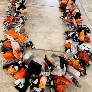 May include: A Halloween-themed garland made of fabric strips in orange, black, white, and gray. The fabric pieces are tied together to create a festive, decorative chain. Some fabric has polka dots, spiderwebs, and ghost patterns.