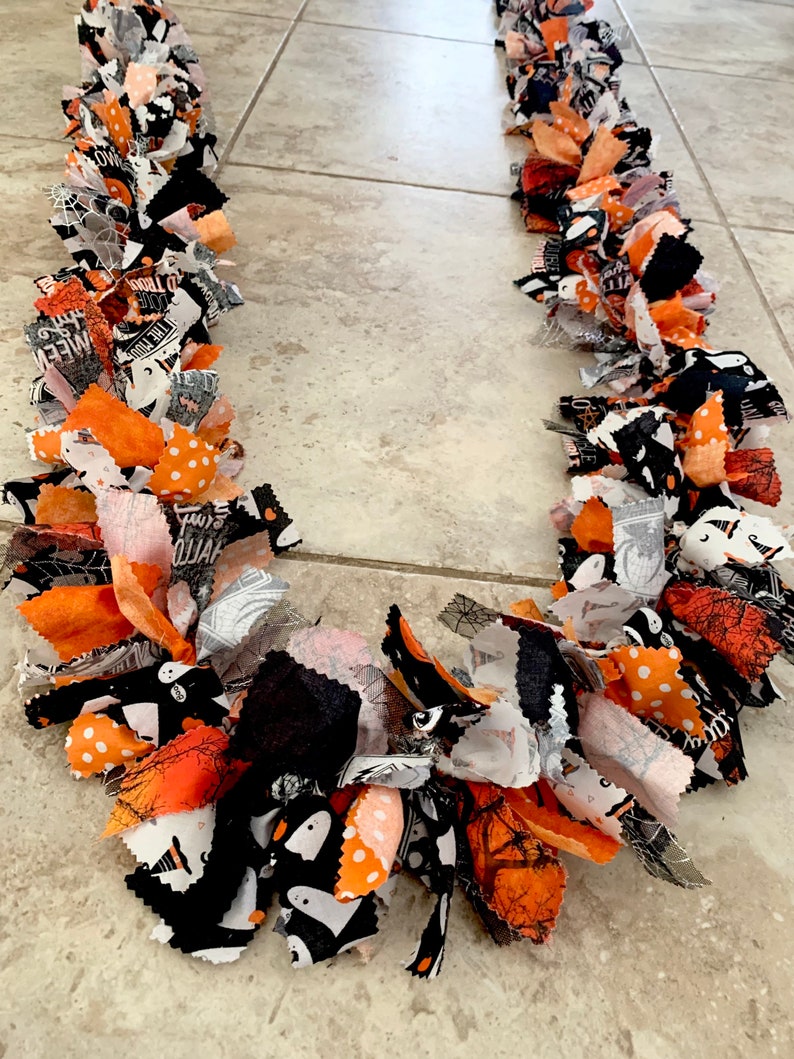 May include: A Halloween-themed garland made of fabric strips in orange, black, white, and gray. The fabric pieces are tied together to create a festive, decorative chain. Some fabric has polka dots, spiderwebs, and ghost patterns.