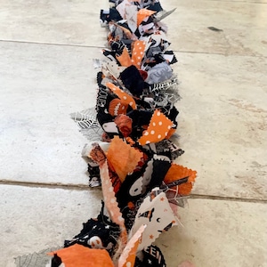 May include: A Halloween-themed fabric garland featuring orange, black, and white patterned fabric strips. The garland includes fabrics with polka dots, ghosts, and spiderwebs. The decorative piece is laid out on a tiled floor.