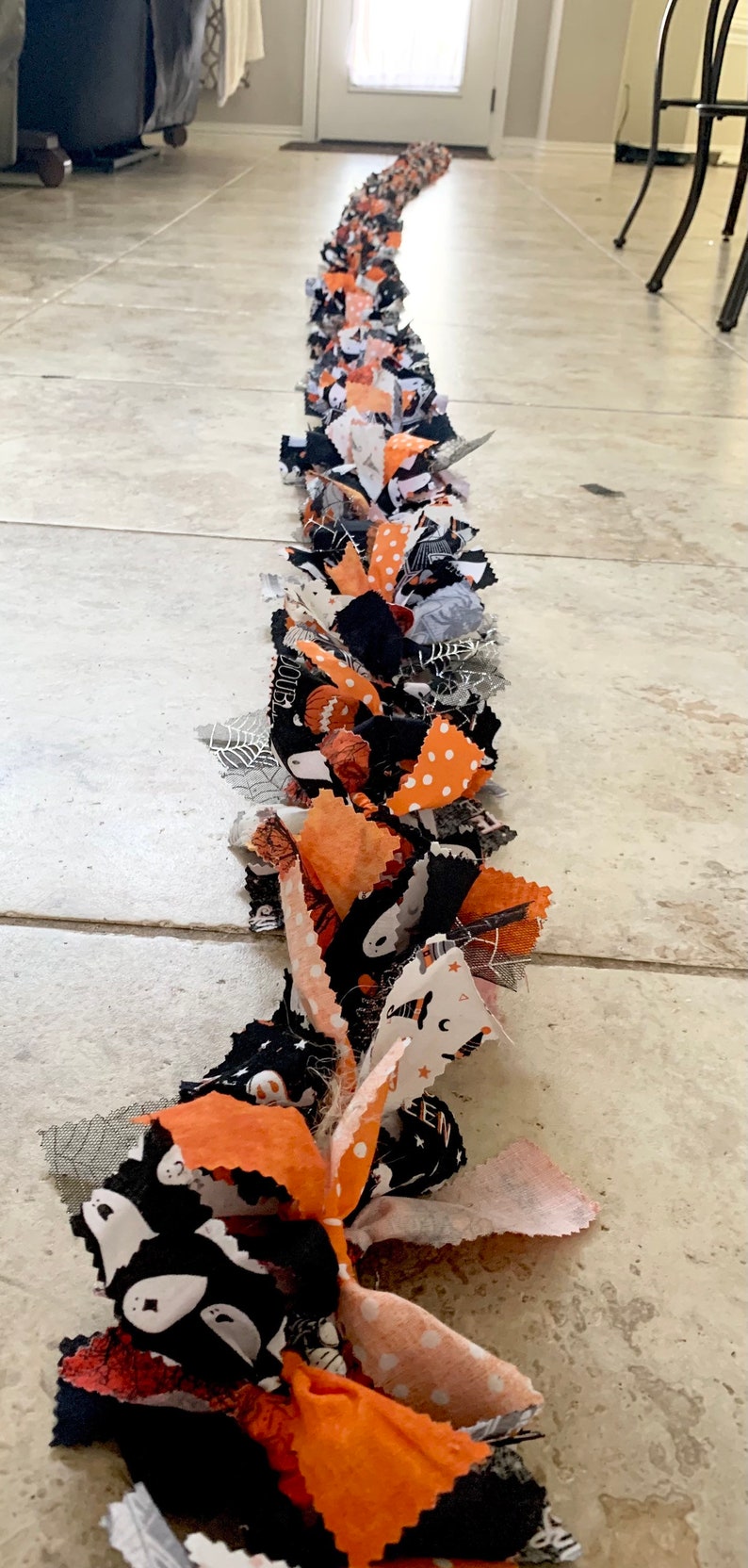 May include: A Halloween-themed fabric garland featuring orange, black, and white patterned fabric strips. The garland includes fabrics with polka dots, ghosts, and spiderwebs. The decorative piece is laid out on a tiled floor.