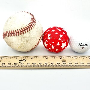 May include: Three balls of different sizes and textures: a baseball, a red ball with white stars, and a white golf ball with the word "Noodle" and the number "2" printed on it. A wooden ruler is placed below the balls.