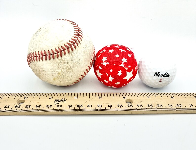 May include: Three balls of different sizes and textures: a baseball, a red ball with white stars, and a white golf ball with the word "Noodle" and the number "2" printed on it. A wooden ruler is placed below the balls.
