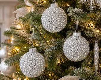 May include: Three spherical Christmas ornaments with a textured, silver glitter finish hanging from thin strings. The ornaments feature a repeating dimpled pattern across their surface and are suspended among green pine branches with warm lights.