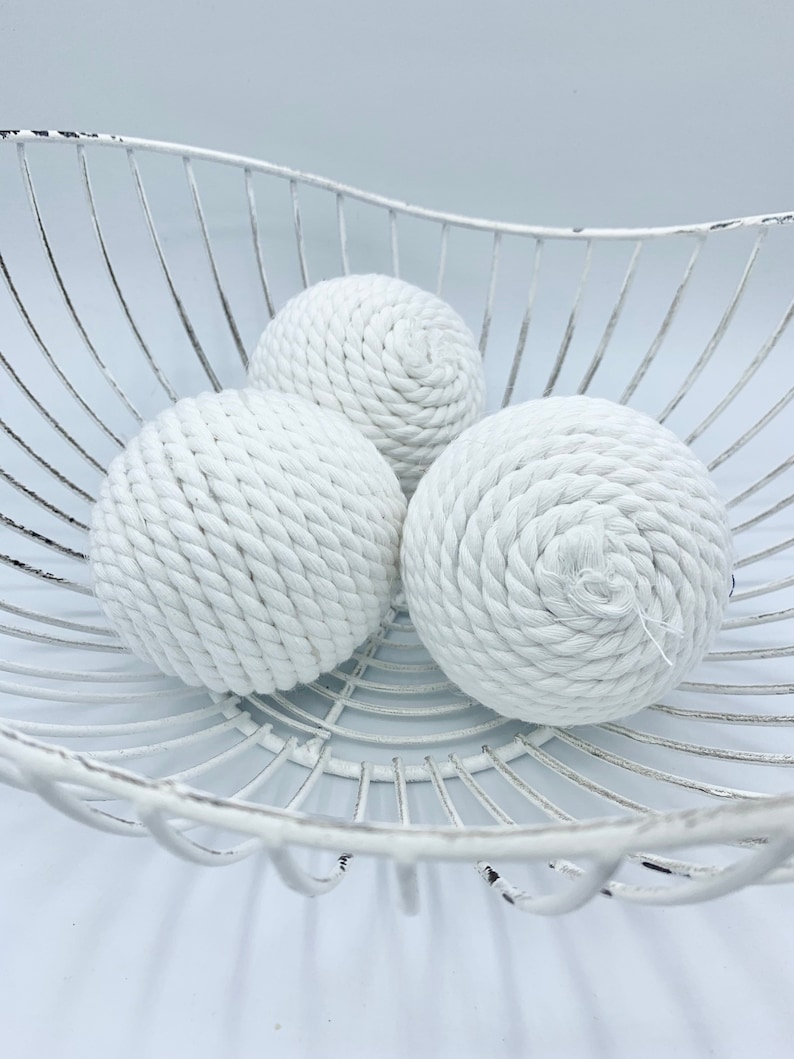 Puede incluir: Tres bolas de cuerda blancas en una cesta de alambre blanca. Las bolas est&aacute;n hechas de cuerda gruesa y tienen un aspecto natural y r&uacute;stico.