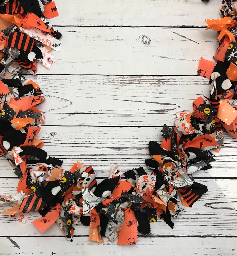 May include: A Halloween-themed wreath made of fabric strips in orange, black, and white. The strips are patterned with bats, spiders, and other spooky designs. The wreath is arranged on a white wooden surface.