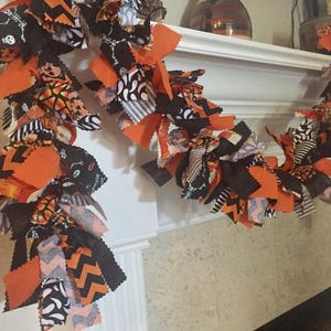 May include: A Halloween garland with orange, black, and white fabric strips. The garland features various patterns, including chevron, skulls, and spiderwebs. It is draped over a white mantelpiece, creating a festive decoration.