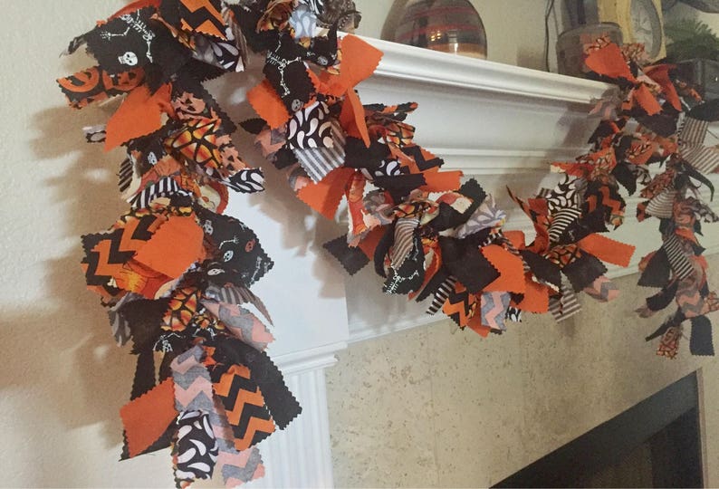 May include: A Halloween garland with orange, black, and white fabric strips. The garland features various patterns, including chevron, skulls, and spiderwebs. It is draped over a white mantelpiece, creating a festive decoration.