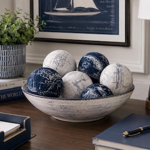 May include: A shallow, distressed white ceramic bowl filled with six decorative spheres. The spheres feature a nautical theme with navy blue and white patterns, including compass roses and map-like illustrations, arranged in a cluster.