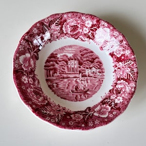 May include: A round, antique-style plate with a red floral border and a central scene depicting a building and figures on a boat. The plate has a scalloped edge and is white in the center.