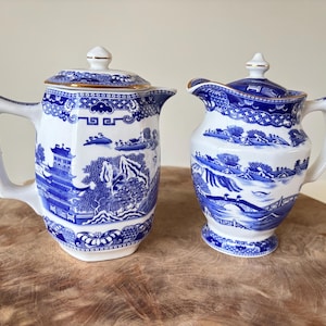 May include: A blue and white ceramic tea set featuring a Willow pattern design. The set comprises a lidded teapot and a creamer. The teapot has a handle and spout, as does the creamer. Both pieces are trimmed with gold detailing.
