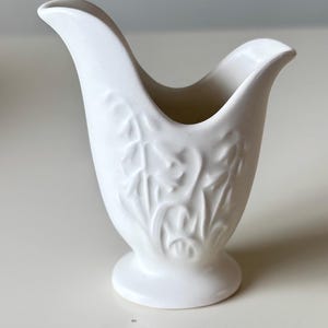 May include: A small, white ceramic vase with a tulip-like shape. The vase features a detailed embossed floral design and a flared rim. The base is a small, circular pedestal. The vase is a solid, matte white color.