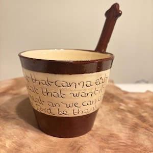 以下が含まれることがあります： 手作りのセラミックマグカップ。外側は濃い茶色、内側はクリーム色です。マグカップには、お揃いの濃い茶色のハンドルが付いており、手書きのテキストが刻まれています。マグカップは木製の表面に置かれています。