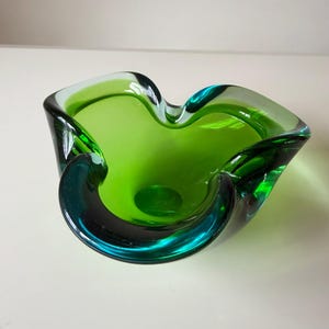 May include: A vibrant green glass bowl featuring a freeform, organic shape with undulating, folded edges. The translucent material displays varying depths of colour, transitioning from a bright lime green centre to deeper emerald tones at the rim.