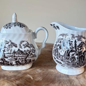 Può includere: Una zuccheriera in ceramica bianca e marrone con coperchio e una lattiera. Entrambi i pezzi presentano un design transferware dettagliato che raffigura una scena rurale. La zuccheriera ha un manico su ogni lato e la lattiera ha un manico curvo.