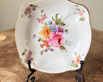 May include: A white, square-shaped porcelain dish with a gold rim, featuring a floral design in pink, yellow, blue, and purple. The dish is displayed on a black metal stand, showcasing its decorative details.