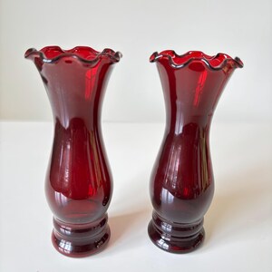 May include: Two ruby red glass vases with frilled rims. The vases have a classic shape, with a flared top and a rounded body. The base of each vase is a darker shade of red. These decorative vases are ideal for displaying flowers.