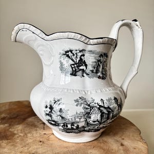 May include: White ceramic pitcher with a black and white pastoral scene. The pitcher has a scalloped rim and a curved handle. The design features figures, trees, and buildings. The pitcher is sitting on a wooden surface.