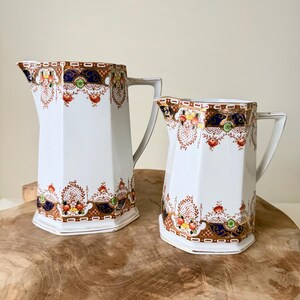 Op de afbeelding: Twee witte keramische kannen met een zeshoekige vorm en een handvat. De kannen zijn voorzien van een decoratief patroon van blauwe, oranje en groene bloemmotieven met goudkleurige accenten langs de boven- en onderrand van elk object.