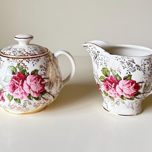May include: A white porcelain sugar bowl with a lid and a creamer. Both pieces are decorated with pink roses, green leaves, and gold filigree. The sugar bowl has two handles and the creamer has a single handle.