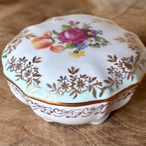 Puede incluir: Pequeña caja de baratijas redonda de porcelana con un diseño floral en la tapa. La tapa presenta un arreglo central de rosas rosas, tulipanes naranjas y otras flores coloridas. Patrones florales dorados y adornos adornan los bordes de la caja.