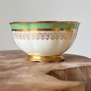 Puede incluir: Un cuenco de cerámica blanca con borde y base dorados, y una banda verde cerca de la parte superior. El cuenco presenta diseños de filigrana dorada ornamentados alrededor del centro. El cuenco está sobre una superficie de madera.