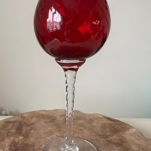 May include: A ruby red glass candle holder with a round bowl-shaped top and a clear, twisted stem. The base is a clear, flat circle. The candle holder sits on a wooden surface.